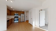This single-floor unit is designed with a clean, contemporary aesthetic that emphasizes open living and functional design. The kitchen is a standout feature, equipped with a full suite of stainless steel appliances including a gas range, built-in microwave, refrigerator, and dishwasher. Warm-toned wood cabinetry extends to the ceiling, providing ample storage, while light countertops and a tile backsplash contribute to a bright, modern feel. Light wood-style flooring runs throughout the main living areas, reflecting natural light and enhancing the sense of openness.

The living area is generously sized and flows seamlessly from the kitchen, creating an adaptable open-plan layout. White walls and recessed lighting maintain a clean, airy atmosphere. A louvered closet door near the entryway adds practical storage, while a separate doorway leads to the bedroom.

The bedroom features neutral-toned carpet for added comfort, a window that brings in natural daylight, and clean white walls offering a calm retreat.

The bathroom is finished to a high standard, featuring large-format floor tiles, a floating wood vanity with an integrated white sink, a backlit mirror, and a full glass-enclosed tub and shower combination with marble-style wall surround. The overall design is cohesive, modern, and move-in ready.
