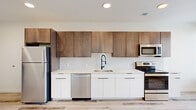 This contemporary studio offers a thoughtfully designed open-plan layout finished with light-toned hardwood flooring throughout, creating a bright and cohesive aesthetic. The kitchen is a standout feature, showcasing a two-tone cabinetry design that pairs warm walnut-finish upper cabinets with sleek white lower cabinets, all set against a classic white subway tile backsplash. The countertops are finished in a light marble-look quartz, providing both durability and elegance. A full suite of stainless steel appliances is included, featuring a refrigerator, electric range, over-the-range microwave, and dishwasher, along with a modern matte black gooseneck faucet at the undermount sink. Recessed lighting is installed throughout the ceiling, ensuring consistent, well-distributed illumination across the entire space. Large floor-to-ceiling windows with horizontal blinds line the living area, inviting abundant natural light and offering open views of the surrounding urban environment. The living area is open and versatile, providing ample room for flexible furniture arrangements. A closet and a private entry door with a keypad lock add to the unit's functionality. The clean white walls and minimalist architectural details contribute to an overall ambiance that is modern, airy, and move-in ready.