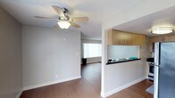 This single-story residence showcases an open and functional floor plan designed to maximize usability. The main living area features warm wood-tone laminate flooring that extends throughout, complementing the neutral, light-toned walls that create a clean and welcoming atmosphere. A five-blade ceiling fan with an integrated light fixture is centrally mounted on the textured ceiling, providing both airflow and ambient lighting.

A defining architectural feature is the pass-through kitchen opening, which connects the kitchen to the living area seamlessly. The kitchen is equipped with warm wood-tone upper cabinetry and dark stone-finish countertops, with a teal accent wall adding a subtle pop of color. The pass-through counter doubles as a breakfast bar or serving surface, enhancing the open feel between spaces.

A window at the far end of the living area allows natural light to filter in through vertical blinds, brightening the interior. The overall design is straightforward and practical, with clean lines, consistent flooring, and a neutral color palette that offers versatility for personalization. The layout flows smoothly from the kitchen area into the living space, making the unit feel cohesive and well-organized.