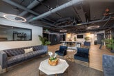 Located at 3638 Motor Ave in Los Angeles, CA, this single-floor amenity and lobby space offers a generously sized, thoughtfully designed environment that blends industrial aesthetics with upscale modern finishes. Throughout the space, exposed black ceilings reveal structural ductwork and piping, while striking circular halo pendant lights provide warm, ambient illumination that contrasts beautifully against the dark overhead palette.

The flooring consists of light wood-tone planks that run continuously through the corridor and lounge areas, lending warmth and cohesion to the space. Wall treatments vary between dark painted surfaces, exposed dark brick, and natural wood-tone panels, creating a layered, urban texture.

The corridor serves as the main circulation spine, lined with Amazon package lockers, potted tropical plants, and a navy upholstered bench alongside an elevator with stainless steel doors. The lounge area transitions into a more refined setting, featuring tufted velvet sofas, gold-accented armchairs, marble-topped coffee tables, a bubble chair, and a communal high-top table with bar stools — ideal for socializing or casual work. Windows along one wall allow natural light to filter in, softening the industrial character. The space is further accented with lush greenery throughout, adding life and vibrancy to the overall ambiance.