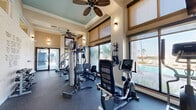 Located at 12001 Belcher Rd S in Pinellas Park, Florida, this fitness center is a well-appointed exercise room designed to support a comprehensive workout experience. The space features elevated ceilings with tray detailing and dual ceiling fans that promote air circulation, contributing to a comfortable workout environment. The warm, neutral-toned walls are complemented by wall sconces that provide soft, ambient lighting throughout.

Large windows with woven roller shades line the perimeter, flooding the room with natural light and offering views of the outdoor pool area and surrounding palm trees — a distinctly Florida ambiance. The durable rubber flooring is practical and well-suited for heavy fitness equipment use.

The room is equipped with a multi-station cable weight machine, a recumbent exercise bike, an elliptical trainer, a treadmill, and an adjustable workout bench, providing options for both strength training and cardiovascular exercise. Wall-mounted resistance bands and additional accessories round out the available equipment. A wall-mounted television is positioned for convenient viewing during cardio sessions. Glass entry doors open directly to the outdoor area, reinforcing the seamless connection between indoor and outdoor amenities. Posted fitness center rules ensure proper use of this shared facility.