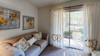 This single-floor residence offers a thoughtfully designed layout that balances comfort and functionality across its key living spaces. The open-concept kitchen is a standout feature, equipped with warm wood cabinetry, white appliances including a refrigerator, electric range, and microwave, as well as a double sink. A built-in breakfast bar with a butcher-block-style countertop and room for multiple barstools provides a casual dining and gathering spot at the heart of the home. The living area is carpeted and bathed in natural light, thanks to a full-length sliding glass door that opens to an outdoor view of mature trees and landscaping, lending the space a calm, connected-to-nature ambiance. The bedroom is generously sized with plush carpet, a ceiling fan with integrated lighting, and a window dressed with sheer curtains that allow soft, diffused light to fill the room. The full bathroom includes a single vanity with a large mirror, a toilet, and a shower-tub combination fitted with a curtain rod. The overall aesthetic throughout is neutral and warm, with beige and cream tones creating a cohesive, inviting atmosphere ideal for everyday living.