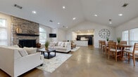 Located at 910 Saint Andrews Drive in Murfreesboro, TN, this single-floor community space offers an open-concept layout that seamlessly connects the living, dining, and kitchen areas. The interior is defined by polished concrete floors throughout, soaring vaulted ceilings with recessed lighting, and crisp white walls that create a bright, welcoming atmosphere.

The living room serves as the focal point of the space, anchored by a floor-to-ceiling stacked stone fireplace flanked by arched windows that flood the room with natural light. Two flat-screen televisions are mounted on either side of the fireplace, adding a functional, modern touch.

The dining area accommodates multiple seating arrangements with high-top tables and chairs, and benefits from a bank of large windows and glass double doors that open directly onto an outdoor patio, offering views of the surrounding community.

The kitchen features dark espresso cabinetry, granite-style countertops, a peninsula island, and a full suite of black appliances including a refrigerator, dishwasher, microwave, and range. A welcoming entry foyer with comfortable seating and decorative accents completes the space, making it ideal for gatherings and community events.