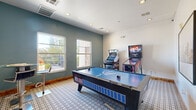 Located at 24770 E Applewood Cir in Aurora, Colorado, this purpose-built game room delivers a compact yet well-appointed entertainment experience. The single-room space is thoughtfully designed with a dual-tone color scheme, pairing a bold slate-blue accent wall with crisp white walls and ceilings to create a lively yet clean atmosphere. Recessed lighting throughout ensures the room is consistently well-lit, complementing the natural light that pours in through two large windows along the accent wall. These windows also offer pleasant views of the surrounding outdoor walkways and landscaping, adding an open, airy quality to the space. The patterned tile flooring anchors the room with a geometric design that adds visual interest underfoot. A full-size air hockey table serves as the centerpiece, while an upright multi-game arcade cabinet and a themed pinball machine line one corner, providing a variety of entertainment options. Two wall-mounted flat-screen televisions are positioned for easy viewing from multiple points in the room. Warm wood baseboards provide a grounding contrast to the contemporary finishes. A high-top table and bar stool offer a comfortable spot to relax between games. The room is accessed through a single interior door, keeping the space self-contained and focused entirely on recreation.