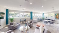 Located at 5100 West Sample Road in Coconut Creek, Florida, this thoughtfully designed community clubhouse offers a welcoming and versatile gathering space for residents. The single-floor layout is anchored by an open-concept lounge area furnished with contemporary grey sofas, accent chairs, and glass-top coffee tables, creating a comfortable setting for relaxation and socializing. A wall-mounted flat-screen television enhances one of the lounge zones, while a dedicated co-working area features a round white pedestal table with modern seating and a built-in desk station, ideal for remote work or study. Throughout the space, bold teal accent columns and wall details provide striking pops of color against a neutral palette of white ceilings and soft grey walls. Patterned carpeting adds texture underfoot, complementing the overall contemporary aesthetic. A row of expansive horizontal windows wraps around the space, flooding the interior with abundant natural light and offering views of the lush, tree-lined surroundings. High-top bistro tables positioned along the window line provide additional casual seating. Recessed lighting illuminates the space evenly, and decorative indoor plants add a refreshing, organic touch. A foyer with vaulted ceilings and restroom access completes this well-appointed community hub.