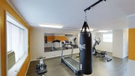 This dedicated fitness space is thoughtfully designed to support a full range of workout routines within a single-level layout. The room features durable hardwood-style flooring that provides both functionality and a clean aesthetic. Large wall-mounted mirrors run along one side of the room, visually expanding the space and allowing users to monitor their form during exercise. A bold "NO EXCUSES" motivational graphic is displayed prominently above the mirrors, setting an energetic tone throughout.

The space is equipped with a variety of cardio and strength training equipment, including a treadmill, an elliptical trainer, a rowing-style machine, and a ceiling-mounted Everlast MMA heavy punching bag — ideal for boxing and martial arts training. Natural light filters in through a window on one side of the room, complementing the bright overhead fluorescent lighting that ensures the space remains well-lit at all times.

The walls feature a neutral color palette with an orange accent panel behind the mirror wall, adding a dynamic visual element that enhances the energy of the environment. The overall layout is open and efficient, allowing for comfortable movement between equipment. This fitness room is well-suited for personal training, cardio sessions, and combat sports practice alike.