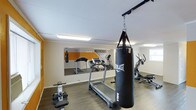 This dedicated fitness space is thoughtfully designed to support a full range of workout routines within a single-level layout. The room features durable hardwood-style flooring that provides both functionality and a clean aesthetic. Large wall-mounted mirrors run along one side of the room, visually expanding the space and allowing users to monitor their form during exercise. A bold "NO EXCUSES" motivational graphic is displayed prominently above the mirrors, setting an energetic tone throughout.

The space is equipped with a variety of cardio and strength training equipment, including a treadmill, an elliptical trainer, a rowing-style machine, and a ceiling-mounted Everlast MMA heavy punching bag — ideal for boxing and martial arts training. Natural light filters in through a window on one side of the room, complementing the bright overhead fluorescent lighting that ensures the space remains well-lit at all times.

The walls feature a neutral color palette with an orange accent panel behind the mirror wall, adding a dynamic visual element that enhances the energy of the environment. The overall layout is open and efficient, allowing for comfortable movement between equipment. This fitness room is well-suited for personal training, cardio sessions, and combat sports practice alike.