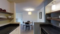 This single-floor residential space offers a functional and well-organized layout with a seamless connection between the kitchen and dining areas. The kitchen is outfitted with white cabinetry, dark countertops, and modern appliances, providing ample workspace and storage. A half-wall partition elegantly separates the kitchen from the dining area, creating a sense of definition while maintaining an open feel. The dining area comfortably accommodates a round table with seating for four, centered beneath a stylish drum pendant light that adds a warm, inviting glow. The space features durable hardwood-style flooring that flows throughout, complementing the neutral white walls and adding a cohesive, contemporary look. A textured ceiling adds a subtle design element overhead. The entry door, visible from the dining area, contributes to the practical flow of the layout. A framed piece of wall art adds a personal touch to the space. Overall, the space presents a clean, move-in-ready environment that balances everyday functionality with a modern, understated aesthetic, making it well-suited for comfortable day-to-day living.