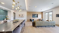 Located at Whistle Stop Lane in Frisco, Texas, this single-story community space offers a thoughtfully designed open-plan layout that seamlessly combines a kitchen and living area. The space is finished with light wood-look flooring throughout, contributing to a clean, contemporary aesthetic that flows from room to room.

The kitchen is a standout feature, anchored by a large dark stone island with seating for multiple guests, illuminated by a series of elegant gold pendant lights. A dramatic floor-to-ceiling teal tile backsplash serves as a bold focal point behind the counters, which are outfitted with white cabinetry, dark stone surfaces, and matte black fixtures. Additional cabinetry and counter space along the perimeter provide ample utility for gatherings and events.

The living area is bright and expansive, benefiting from large windows along multiple walls that flood the space with natural light and frame panoramic views of the surrounding Frisco landscape. Full-length glass-panel doors open to a balcony, further extending the connection to the outdoors. Recessed ceiling lighting complements the natural light throughout.

A separate nook within the space accommodates recreational use, currently featuring an arcade game station set on protective mat flooring. The overall ambiance is modern and inviting, well-suited for social gatherings and community activities.