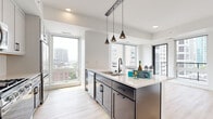 This contemporary urban residence is thoughtfully designed with a clean, modern aesthetic and high-quality finishes throughout. The open-concept kitchen serves as the heart of the home, featuring a large center island with a white quartz countertop, dark cabinetry with matte black hardware, and a suite of stainless steel appliances including a refrigerator, microwave, and gas range. White subway tile backsplash and white upper cabinetry add a bright contrast to the sleek lower cabinets. Pendant lighting in a matte black finish hangs above the island, complementing the overall design palette. Light wood-tone flooring flows through the kitchen and living areas, adding warmth to the crisp, white interior.

Floor-to-ceiling windows throughout the residence flood the space with abundant natural light and frame expansive city skyline views, highlighting the elevated position of the unit. The bedroom is generously proportioned, finished with plush carpeting and a large window that continues the panoramic urban vistas. The bathroom offers a refined, spa-like atmosphere with a dual-sink white quartz vanity, matte black fixtures, a large mirror, and a glass-enclosed walk-in shower with white tile surround. Every detail of this residence reflects a commitment to modern, urban living.