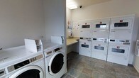 This shared laundry facility is a practical, well-organized space designed to accommodate multiple users efficiently. The room features a lineup of commercial-grade front-loading washing machines along one side, each clearly numbered for easy identification. Opposite and along the back wall, stacked dryer units—also individually numbered—provide ample drying capacity, with their large porthole windows allowing users to monitor cycles at a glance. A built-in folding counter runs along the back wall, offering a convenient surface for sorting and folding laundry. The room is finished with neutral-toned walls and durable slate-pattern tile flooring, contributing to a clean and functional aesthetic. Overhead fluorescent lighting ensures the space is well-lit, while an overhead security camera provides added safety and oversight. A dispenser mounted on the wall offers accessible supplies, and informational notices are posted near the machines. The layout is thoughtfully arranged to allow comfortable movement between machines, making the overall experience of using the facility straightforward and efficient.