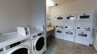 This shared laundry facility is a practical, well-organized space designed to accommodate multiple users efficiently. The room features a lineup of commercial-grade front-loading washing machines along one side, each clearly numbered for easy identification. Opposite and along the back wall, stacked dryer units—also individually numbered—provide ample drying capacity, with their large porthole windows allowing users to monitor cycles at a glance. A built-in folding counter runs along the back wall, offering a convenient surface for sorting and folding laundry. The room is finished with neutral-toned walls and durable slate-pattern tile flooring, contributing to a clean and functional aesthetic. Overhead fluorescent lighting ensures the space is well-lit, while an overhead security camera provides added safety and oversight. A dispenser mounted on the wall offers accessible supplies, and informational notices are posted near the machines. The layout is thoughtfully arranged to allow comfortable movement between machines, making the overall experience of using the facility straightforward and efficient.