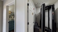 This single-floor space offers a well-organized and intimate layout designed for comfort and practicality. The entryway sets the tone with classic six-panel white doors, accented by matte black hinges and hardware that add a modern contrast to the traditional styling. A textured ceiling adds architectural character throughout, while recessed vents ensure efficient climate control. Safety is addressed with a clearly visible fire alarm system near the entry. Just beyond the door, the space transitions into a more personal area where a sleek, dark upholstered bench provides a convenient spot for seating or storage at the entry point. A tall, modern black beverage or wine cooler stands nearby, contributing both functionality and a contemporary aesthetic. To one side, a glimpse into an adjacent room reveals a teal-painted dresser adorned with a floral arrangement, introducing a pop of color and a welcoming, homey ambiance. The combination of neutral wall tones, classic millwork, and carefully chosen furnishings creates a space that balances everyday practicality with inviting character. Overall, the space presents a thoughtfully arranged environment that feels both functional and comfortable.
