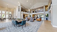 Located at 9246 Highland Creek Pkwy in Charlotte, NC, this single-floor community clubhouse is designed to serve as a central gathering hub for residents. The grand entrance lobby immediately impresses with soaring double-height ceilings, stately columns, and an elegant chandelier overhead. Light wood-look flooring flows throughout, complemented by neutral warm tones and carefully curated artwork and decorative accents on the walls.

The main lounge area offers a generous arrangement of mixed seating, including upholstered armchairs and sofas, alongside a formal dining table with teal accent chairs — all set atop a large patterned area rug. Expansive floor-to-ceiling windows along one wall flood the space with natural light while providing views of the outdoor pool and surrounding community grounds.

A well-appointed community kitchen features a large island with seating, white upper cabinetry, patterned tile backsplash, and a dedicated coffee and beverage station with grey lower cabinets and decorative mirror wall art. An adjacent open dining area with multiple tables and modern chandelier lighting serves as a casual gathering and dining space.

A private conference and media room includes a round table, task chairs, built-in shelving, and a large wall-mounted display screen, suitable for meetings or group activities. Decorative touches such as tall potted plants, triptych artwork, and geometric wall installations add warmth and character throughout.