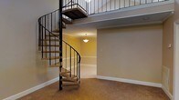 This space is defined by a dramatic spiral staircase that serves as its architectural centerpiece, constructed with carpeted treads and a sleek wrought iron railing that curves gracefully from the main level up to an open upper loft area. The loft overlooks the main floor and is enclosed by matching wrought iron baluster railings, reinforcing the cohesive design language throughout the space.

The main level features a combination of wall-to-wall carpet and tile flooring, transitioning between distinct areas. Neutral beige tones coat the walls throughout, complemented by clean white baseboards and crown molding that add a polished, finished look. A pendant light fixture hangs from the ceiling in the tiled area visible beyond the staircase, providing warm, focused illumination and defining that zone as a separate functional space.

The overall layout is open and airy, with the upper loft contributing to a sense of vertical volume and spaciousness. The design balances practicality with visual interest, making the spiral staircase both a functional connector between levels and a standout decorative element within the space.