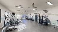 Located at 20401 North 19th Avenue in Phoenix, AZ, this fitness center offers a generously sized, single-floor exercise space designed to support a comprehensive workout experience. The room is bright and airy, benefiting from abundant natural light streaming through large windows that offer views of the surrounding neighborhood. Multiple ceiling fans and fluorescent lighting fixtures keep the environment comfortable and well-illuminated throughout.

The exercise area is divided into distinct functional zones. The cardio section features a row of treadmills positioned along a windowed wall, accompanied by a wall-mounted flat-screen TV for entertainment during workouts. Elliptical trainers and other cardio equipment are also available. The strength training area is well-stocked with cable machines, a multi-station strength unit, a dumbbell rack, medicine balls, and a balance board for functional training.

The interior design is clean and modern, with white walls accented by a soft gray horizontal band. A striking large-scale mural of an athletic figure adorns one wall, adding a dynamic, motivational aesthetic to the space. Durable rubberized flooring covers the main workout area, with tile flooring near the entrance. The layout flows efficiently, providing ample open floor space for movement and stretching. Shelving units offer organized storage for accessories, completing a polished and fully functional fitness facility.