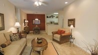 This single-floor residence offers an open-concept layout that seamlessly connects the living room, dining area, and kitchen. The living room is furnished with plush, neutral-toned seating accented by warm red throw pillows, centered around a decorative round wooden coffee table. Recessed lighting and a ceiling fan overhead provide both functionality and comfort. Soft, wall-to-wall carpeting throughout the main living and dining areas contributes to a warm, inviting atmosphere. A bold, deep-red accent wall in the dining area serves as a striking focal point, complementing the warm beige tones found throughout the space. The dining area accommodates a wooden table and chairs beneath an elegant chandelier, making it well-suited for intimate gatherings. Adjacent to the dining space, the kitchen features a pass-through opening with a raised counter, white cabinetry, and under-cabinet lighting, creating a bright and functional workspace. Decorative touches such as framed artwork and table lamps add personality throughout the space, while the cohesive color palette of warm earth tones and neutral walls creates a unified, welcoming ambiance.