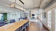 Located at Avalon Gardens in Nanuet, NY, this single-floor communal amenity space is thoughtfully designed to serve as a gathering and lounge destination for residents. The open-plan layout flows seamlessly from a well-appointed galley kitchen through a dining area and into a spacious lounge, all unified by rich, dark hardwood flooring throughout.

The kitchen features light wood cabinetry, gray countertops, marble-style tile backsplash, stainless steel appliances, and large-format tile flooring — offering a functional and clean workspace for hosting events. Adjacent to the kitchen, a long natural wood communal dining table anchors the dining zone beneath an industrial-style rectangular pendant light, with ample seating for group gatherings.

The lounge area is the architectural centerpiece of the space, boasting soaring double-height ceilings, a dramatic floor-to-ceiling whitewashed stacked stone fireplace with a mounted flat-screen television, and a generous seating arrangement. A wall of French doors with arched transom windows floods the space with natural light and provides direct access to an outdoor deck. A glass-partitioned room adjacent to the main area offers a more private seating nook. The overall ambiance is modern yet welcoming, with a cool blue-gray color palette and carefully curated furnishings.