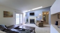 This single-floor residence offers a welcoming and well-appointed living environment designed for comfort and ease. The living room serves as the heart of the space, showcasing a vaulted ceiling with skylights that flood the room with abundant natural light throughout the day. A tiled fireplace with a classic white mantel anchors the room, creating a cozy focal point complemented by soft blue accent walls that lend a calm, airy atmosphere. Track lighting along the skylight shaft adds flexibility for evening ambiance. French doors with divided-light panes open to the outdoors, further enhancing the sense of openness and providing a seamless connection to the exterior. Plush wall-to-wall carpeting in a neutral tone runs underfoot, adding warmth and comfort to the space. The living area is generously sized, offering ample room for seating arrangements centered around both the fireplace and a wall-mounted television. A hallway visible from the main living area leads to additional rooms, hinting at a thoughtfully laid-out floor plan. Overall, this space balances practical design with inviting details, making it a comfortable and functional home.