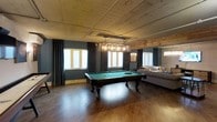 This single-floor space blends rustic character with modern comfort, creating an inviting atmosphere ideal for entertainment and relaxation. The centerpiece of the main area is a full-size pool table with a green felt surface, positioned beneath a striking industrial-style pendant light fixture adorned with exposed Edison bulbs. The warm, rich hardwood flooring grounds the space and complements the natural wood-paneled walls and a weathered, whitewashed plank ceiling that adds a distinctive rustic charm. Deep teal-toned walls are accented by elegant dark curtains framing multiple windows that allow generous natural light to filter throughout. A cozy lounge area is tucked along one side of the room, furnished with a plush sofa and soft furnishings, offering a comfortable retreat from the game area. The open layout provides a generous sense of space and flow between the entertainment and relaxation zones. Additional details, such as a pinball or arcade machine visible along one wall, further enhance the recreational character of the space. The overall ambiance is warm and welcoming, combining industrial and rustic design elements to create a space that is both visually striking and functionally versatile.