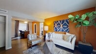 This single-floor residence showcases a thoughtfully designed interior that balances bold color choices with contemporary finishes throughout. The living area is immediately striking, defined by a rich amber-orange accent wall that serves as a dramatic backdrop for a tufted white sofa, metallic accent chairs, and a geometric stone coffee table. Dark hardwood-style flooring runs consistently throughout the space, grounding the vibrant color palette. A large indoor plant and curated artwork add visual depth to the room, while natural light filters in through windows dressed with layered curtains and blinds.

The kitchen is well-equipped and features dark gray cabinetry, white countertops, a double-basin stainless steel sink, a gas range, and an eye-catching mosaic tile backsplash in cool blue and gray tones that extends across the full cooking area.

The bedroom continues the bold amber accent wall theme, complemented by a ceiling fan, large tufted upholstered bed frame, and windows with privacy blinds and drapes that allow for natural light control.

Two bathrooms offer dark-toned vanity cabinetry with white countertops, framed mirrors, and coordinating mosaic tile accents, presenting a clean and modern aesthetic consistent with the overall design of the space.