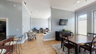Located at 2500 Grove Club Ln in Cary, NC, this single-floor community space offers a thoughtfully designed layout that blends comfort and functionality. Throughout the space, warm hardwood floors and cool gray walls create a cohesive, inviting atmosphere, complemented by crown molding and recessed lighting.

The main lounge area is a standout, featuring soaring ceilings, large windows that flood the room with natural light, and a stone-accented fireplace set within an elegant arched alcove with a wall-mounted TV above. A leather armchair and ottoman provide a relaxed seating option in this cozy corner.

Adjacent to the lounge is a dining and media area with a dark wood dining table and chairs, a flat-screen TV mounted on the wall, and additional seating. This space flows seamlessly into a business center equipped with multiple computer workstations, offering a productive environment for residents.

A curved granite-topped bar with rich wood paneling and bar stool seating serves as an attractive focal point and social hub. The kitchenette nearby features dark cabinetry extending to the ceiling, dark granite countertops, a stainless steel sink, and ample storage — ideal for catering or everyday use. The overall design balances elegant architectural details with practical amenities throughout.
