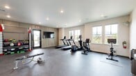 Located at 1800 NW 115th Terr in Oklahoma City, Oklahoma, this fitness center offers a comprehensive and well-organized exercise space designed to support a variety of workout routines. The single-floor layout is finished with durable rubber flooring throughout, providing a safe and practical surface for all types of training activities.

The cardio zone is prominently featured and includes multiple treadmills, an elliptical trainer, and a stationary bike, all positioned to take advantage of the natural light streaming in through multiple large windows along the exterior wall. These windows also provide views of the surrounding outdoor greenery, contributing to an open and energizing atmosphere.

A dedicated strength and functional training area houses a multi-station cable machine, an adjustable weight bench, a full dumbbell rack, medicine balls, resistance bands, and additional free weight equipment — offering options for both beginners and experienced athletes. A wall-mounted mirror in the strength area enhances spatial depth and aids in form monitoring during workouts.

The room also features built-in cabinetry along one wall, a wall-mounted flat-screen television, and recessed lighting overhead for a clean, modern aesthetic. A water fountain is conveniently located within the space. Direct exterior door access adds ease of entry and exit. The overall ambiance is bright, clean, and purposefully designed for focused physical training.