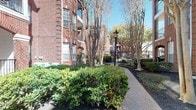 This single-floor residence is situated within a handsome multi-story brick building, surrounded by thoughtfully landscaped grounds. The exterior environment features a winding pathway lined with manicured hedgerows, mature crepe myrtle trees, and classic black lamp posts, creating a refined and welcoming approach to the building.

Inside, the interior reflects a contemporary aesthetic grounded in quality materials and understated design. The dining area serves as a notable focal point, centered around a substantial round green marble table accompanied by woven rattan chairs atop a natural fiber rug. Polished concrete floors extend throughout, lending an industrial-modern character to the space while providing visual continuity between rooms.

The walls are finished in a clean, neutral palette, accented by large-scale framed artwork that adds artistic depth. A striking sliding barn door with a black metal track provides a stylish and functional room divider, connecting the dining area to adjacent spaces. Recessed lighting illuminates the interiors efficiently, complementing the natural light that filters in through the windows. Textured wall finishes add subtle warmth and dimension throughout.

The overall ambiance is sophisticated and thoughtfully curated, balancing industrial elements with organic textures to create an inviting and visually interesting living environment.