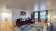 This single-floor residence offers a thoughtfully arranged layout that makes efficient use of its space. The living room serves as the centerpiece of the unit, showcasing warm hardwood flooring that extends throughout the main areas. Large windows dressed with bold teal grommet curtains flood the room with natural light and offer views of the surrounding neighborhood. A spacious sectional sofa anchors the space, complemented by a vibrant multicolored area rug that adds a lively touch to the neutral walls. Abstract artwork displayed above the sofa adds a decorative focal point, while a ladder-style shelving unit provides additional storage and style.

The kitchen is compact yet functional, featuring warm-toned wooden cabinetry that extends to the ceiling for ample storage. It is equipped with a gas range, a full-size refrigerator, and a double-basin sink with dark countertops. Tile flooring in the kitchen area provides a practical transition from the hardwood found elsewhere.

The bedroom is a calm retreat, with hardwood floors continuing underfoot and a window framed by cheerful yellow curtains that let in soft, natural light. The room offers adequate space for a full bedroom set, including matching nightstands and a dresser, providing comfortable and organized living.