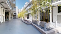 This contemporary residential common area is thoughtfully designed to offer both functionality and aesthetic appeal. At its heart is a beautifully landscaped open-air courtyard, where tall bamboo clusters rise from concrete planters filled with decorative river rock. The smooth, light-toned ground surface extends throughout the corridor, complementing the clean lines of the surrounding multi-story building facades adorned with balconies and large windows that overlook the shared space. Abundant natural light fills the courtyard, creating an inviting and tranquil atmosphere.

Accessible and practical, the space includes a commercial-grade elevator with stainless steel doors, conveniently located alongside a secondary access door — ensuring smooth movement throughout the building. A dedicated mailroom area houses an organized array of individual mailboxes alongside a modern package locker system, providing secure and efficient parcel management for all residents. The consistent use of neutral tones, concrete finishes, and structured landscaping throughout the common areas reflects a cohesive, contemporary architectural design focused on both utility and livability.
