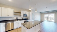 This single-floor unit offers a thoughtfully designed layout across a comfortable footprint, accommodating two bedrooms, two bathrooms, two closets, and a hallway. The open-concept kitchen and living area serves as the centerpiece of the space, featuring white shaker-style cabinetry, dark quartz countertops, a central island with pendant lighting overhead, and a full suite of stainless steel appliances including a refrigerator, range, microwave, and dishwasher. Light wood-look flooring runs throughout the main living areas, complementing the clean, contemporary aesthetic. The living area extends to a private balcony through sliding glass doors, offering outdoor views and natural light. An exposed ductwork element adds an industrial accent to the living space. The two bedrooms are carpeted and include windows with blinds; the primary bedroom offers a dedicated closet and connects to a bathroom featuring white cabinetry, dark quartz countertops, a large vanity mirror, and black hardware accents. The second bathroom includes a tub-shower combination, hardwood-style flooring, and matching finishes, maintaining a consistent modern design throughout the unit.