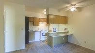 This single-floor residence offers a functional and practical layout centered around an open-concept kitchen and living area. The kitchen is well-appointed with warm wood-toned cabinetry spanning both upper and lower sections, providing ample storage space. White appliances, including a range, dishwasher, and refrigerator, complement the clean, light-colored walls. A peninsula counter extends into the living space, creating a natural divider between the kitchen and the adjacent carpeted living area while also offering additional prep and casual dining space. The kitchen area features tile flooring that transitions seamlessly into carpeted living space, adding a sense of delineation between functional zones. A ceiling fan with a light fixture is positioned overhead, ensuring comfortable air circulation throughout the open area. The textured ceiling adds a subtle architectural detail consistent throughout the space. The overall layout is designed with everyday practicality in mind, offering a cohesive flow between the kitchen and living areas. The neutral color palette of warm beige tones and light walls creates a welcoming and comfortable atmosphere throughout the home.
