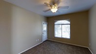 This single-floor space offers a practical and comfortable layout designed with everyday living in mind. The interior is characterized by warm, neutral-toned walls that create a calm and welcoming ambiance throughout. The main living area features wall-to-wall carpeting that adds softness underfoot, while white baseboards provide a clean, finished look along the perimeter.

Natural light flows generously into the space through a large window fitted with horizontal blinds, allowing for adjustable light control. A glazed exterior door further brightens the room while providing convenient access to the outdoors. The view through both the window and door suggests a lightly landscaped outdoor setting with mature trees nearby.

Overhead, a multi-blade ceiling fan with an integrated light fixture serves both as a functional lighting source and an aid in maintaining air circulation, contributing to the overall comfort of the space. The textured ceiling adds a subtle architectural detail to the room.

The overall design is straightforward and functional, emphasizing comfort and practicality. The combination of carpeted flooring, neutral walls, and well-placed windows ensures the space feels bright and livable, making it well-suited for a variety of uses.