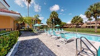 Located at Midori in Tamarac, Florida, this outdoor amenity area is designed to deliver a resort-style experience in a single-story layout. The centerpiece is a generously sized swimming pool with crystal-clear blue water, accessible via stainless steel entry rails and framed by a broad paver-stone deck. Rows of turquoise lounge chairs are arranged along the poolside, offering ample space for sunbathing and relaxation under the South Florida sun. Shaded seating areas with teal umbrellas and bistro tables provide a comfortable retreat from the heat. Lush, mature palm trees surround the pool area, enhancing the tropical ambiance while offering natural shade and scenic views of the landscaped grounds beyond the fenced perimeter. A covered poolside building, finished in a warm golden-yellow stucco exterior with bold blue accents, houses restroom and locker facilities, posted pool rules, and emergency equipment. A tiled walkway corridor runs alongside the building, incorporating decorative planters with mature palms rooted in the pavement. The overall atmosphere is bright, open, and inviting, reflecting a well-maintained community space ideal for outdoor leisure and social gatherings in a classic South Florida setting.