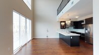 This contemporary two-story space makes an immediate impression with its dramatic double-height ceilings that flood the open-concept living area with an expansive sense of volume and light. Large windows dressed with horizontal blinds invite natural light to pour across the rich hardwood floors, enhancing the warm yet modern aesthetic throughout.

The main level seamlessly connects the living area to a well-appointed kitchen, where dark cabinetry is paired with crisp white countertops for a striking contrast. A generous kitchen island with an integrated sink serves as both a functional workspace and a social centerpiece. Above, a pair of glamorous crystal-style pendant chandeliers adds a refined, eye-catching touch. A stainless steel range hood and glass tile backsplash further elevate the kitchen's polished design.

An upper-level mezzanine overlooks the main living space via a sleek metal railing, offering an open connection between floors while maintaining an airy, loft-inspired feel. This thoughtful architectural detail reinforces the sense of verticality and openness that defines the space. The neutral color palette, clean lines, and carefully chosen finishes throughout create a cohesive and inviting environment suited to both everyday living and entertaining.