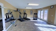 This single-floor fitness room is designed to support a well-rounded cardio workout in a clean, comfortable environment. The space is outfitted with several key pieces of cardio equipment, including an elliptical trainer, a treadmill, and an additional standalone cardio machine, all arranged to allow ample room for movement between stations. Free weights are also available on the floor, adding to the versatility of the workout options. The room features warm, neutral-toned walls and durable stone-pattern flooring that is both practical and easy to maintain. Overhead fluorescent lighting ensures the space is well-lit throughout, while a set of elegant French doors along one wall floods the room with natural light and provides a view of the outdoors, creating an inviting atmosphere. A wall-mounted television is positioned for viewing during workouts, enhancing the overall exercise experience. Framed artwork on the walls adds a subtle decorative touch, balancing the functional nature of the space with a sense of comfort. The exit signage and safety features are clearly visible, reflecting a thoughtfully planned and safety-conscious layout. Overall, this fitness room offers a practical and welcoming environment for daily exercise.