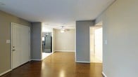 This single-floor residence offers a thoughtfully arranged open-concept layout that promotes a sense of spaciousness and easy flow between living areas. The main living space features rich, warm-toned hardwood flooring that extends throughout, creating a cohesive and inviting atmosphere. A ceiling fan with integrated lighting is centrally positioned overhead, providing both comfort and ambient illumination.

The living area transitions seamlessly into the kitchen, which is equipped with dark wood cabinetry and black appliances, including a full-size refrigerator and range. The contrast between the darker kitchen finishes and the neutral gray-toned walls adds visual depth to the open floor plan.

The walls throughout are finished in a cool, medium gray tone complemented by crisp white baseboards and trim, lending the space a clean and modern aesthetic. A hallway visible from the main living area leads to additional rooms, with a door opening to a bright, well-lit space — likely a bedroom or bathroom — that adds to the overall sense of connectivity within the home. The layout is practical and versatile, offering a functional single-level living experience with consistent finishes throughout.