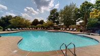 Located at 1113 Powers Ferry Pl SE in Marietta, Georgia, this outdoor amenity space centers around a generously sized freeform swimming pool with crystal-clear turquoise water. The pool's organic, curved shape lends a resort-like character to the area, complemented by stainless steel entry rails for convenient access. Surrounding the pool is a broad, stone-paved sun deck finished in warm earth tones, offering ample space for relaxation and recreation. Rows of modern lounge chairs line the perimeter of the pool deck, providing abundant seating for sunbathing and leisure. A shaded umbrella station offers a respite from the sun, adding to the functionality of the space. The pool area is framed by mature trees and well-maintained greenery, including trimmed shrubs and landscaped lawn areas that create a natural, private atmosphere. A decorative rock feature accents one side of the pool, adding a natural design element. In the background, a handsome brick and wood-sided clubhouse with a stone chimney and pitched roof overlooks the pool, reinforcing the warm, community-oriented character of the space. The surrounding canopy of tall trees provides shade along the edges, enhancing the serene, secluded ambiance of this outdoor retreat.