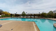 Located at 1245 E Prairie Brook Dr in Palatine, Illinois, this outdoor community pool facility offers a well-appointed recreational space designed for relaxation and leisure. The centerpiece is a generously sized swimming pool with clear blue water, surrounded by a wide concrete sundeck that provides ample room for lounge chairs and poolside gatherings. Ornamental lamp posts line the perimeter, adding a classic aesthetic to the outdoor environment. A decorative pergola structure anchors one end of the pool area, while a covered gazebo with picnic-style seating offers a shaded retreat nearby. Outdoor tables and chairs are positioned along the deck, creating comfortable spots for socializing. The facility is enclosed by a black iron fence that frames the space while preserving open sightlines to a scenic pond and surrounding tree line beyond, providing a tranquil natural backdrop. The adjacent single-story building features classic brick-and-siding construction with large windows that overlook the pool, and a direct entry door provides convenient access between the indoor and outdoor areas. The combination of thoughtful landscaping, waterfront views, and quality amenities makes this a standout community recreational destination.