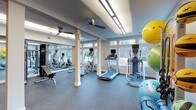 This fitness center offers a thoughtfully designed workout space that balances functionality with a welcoming atmosphere. Large windows line the walls, flooding the interior with natural light and providing views of the surrounding wooded landscape, creating an energizing environment for exercise. The ceiling features recessed linear lighting alongside ceiling fans, ensuring comfortable airflow and consistent illumination throughout the space.

The flooring consists of durable, cushioned gray rubber material, ideal for both cardio and strength training activities. Structural columns with classic white trim are evenly spaced throughout the room, adding an architectural character while defining different workout zones. A mirrored wall on one side visually expands the space and supports proper form during exercise.

The fitness center is outfitted with a variety of cardio equipment, including treadmills, an elliptical trainer, and stationary bikes, each equipped with individual monitors for entertainment or workout tracking. A dedicated strength training area includes a weight bench and multi-use cable machine. Stability and exercise balls in multiple sizes round out the functional equipment offerings. The overall ambiance is bright, clean, and motivating.