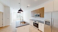 Located at 5111 Abercorn St in Savannah, Georgia, this single-floor unit offers a thoughtfully designed, contemporary living space. The open-concept kitchen and living area serves as the heart of the unit, featuring light hardwood-style flooring throughout. The kitchen is outfitted with stainless steel appliances — including a side-by-side refrigerator, electric range, and built-in microwave — complemented by a two-tone cabinetry design that pairs warm wood upper cabinets with crisp white lower cabinets. A subway tile backsplash and dark granite countertops add a polished finish, while a central island with an undermount sink and striking matte black pendant lighting anchors the space. Recessed ceiling lights provide additional illumination throughout. The bedroom features soft carpeting, a ceiling fan, and a window with outdoor views, creating a comfortable and well-lit retreat. A spacious walk-in closet with wire shelving offers generous storage. The bathroom includes a granite vanity, tiled tub-shower combination, and a window that brings in natural light. The unit's neutral white walls and clean architectural lines create a bright, modern ambiance throughout.