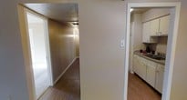 This single-floor residence offers a straightforward and functional layout designed for comfortable everyday living. A central hallway serves as the spine of the home, connecting the various living spaces and providing a clear sense of flow throughout. The hallway is finished with warm hardwood-style flooring that extends seamlessly into the adjacent rooms, lending a cohesive aesthetic to the interior.

The kitchen is visible from the main corridor and features white cabinetry that lines the walls, offering generous storage space. A countertop with a built-in sink adds practicality, and the cabinetry's clean, light finish keeps the space feeling open and organized.

The walls throughout are finished in neutral tones, complementing the warm flooring and contributing to an understated, welcoming ambiance. Textured ceilings add a subtle detail to the overhead surfaces. Natural light appears to filter in through doorways and openings, brightening the interior spaces.

Overall, this residence presents a no-frills, well-connected layout with consistent finishes throughout, making it a practical and livable single-floor space suited for a variety of occupants.