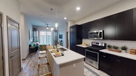 This contemporary single-floor residence offers a thoughtfully designed open-concept layout that seamlessly connects the kitchen and living areas. The kitchen is a standout feature, showcasing bold dark cabinetry that extends floor to ceiling, contrasted by bright white countertops and a classic subway tile backsplash. A large center island with seating provides an ideal space for casual dining, while stainless steel appliances — including a gas range, built-in microwave, and side-by-side refrigerator — complete the chef-ready setup. Elegant pendant lighting illuminates the island, adding a refined touch to the space.

The open living area is bathed in natural light from a sliding glass door that leads to an outdoor balcony, with a ceiling fan overhead adding to the comfort of the room. Light wood-toned flooring runs throughout the main living spaces, enhancing the modern aesthetic.

The residence includes two well-proportioned bedrooms, each featuring plush carpeting, large windows with outdoor views, and ceiling fans. One bedroom is served by a private en-suite bathroom with light wood-style flooring, a dark vanity, and tiled walls, alongside a spacious walk-in closet with built-in shelving and hanging rods — offering generous storage and organization options.