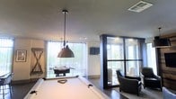 This contemporary recreational lounge is thoughtfully designed to offer both entertainment and relaxation in a single, open-plan space. The room is anchored by a full-sized pool table positioned beneath a striking industrial-style dome pendant light, which serves as a bold focal point against the smooth, neutral-toned ceiling. A foosball table adds to the entertainment options near the expansive windows, which are dressed with sheer vertical blinds that filter generous amounts of natural light throughout the space. Dark hardwood flooring provides a rich contrast to the lighter walls and furnishings, lending the room a polished, modern aesthetic. A partially visible seating area, furnished with sleek dark armchairs, is framed by a bold dark-finished structural column and a floor-to-ceiling glass partition, creating a defined yet open lounge nook. A wall-mounted flat-screen display is also present, adding a media dimension to the space. The abundance of large windows along multiple walls ensures the interior remains bright and connected to the outdoors, with views of surrounding greenery visible from within. Overall, the space strikes a balance between lively entertainment and comfortable lounging in a well-appointed, contemporary setting.