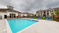 Located at 1 Kerry Park Cir in Delaware, Ohio, this outdoor amenity space centers around a generously sized swimming pool with vivid blue water, offering a welcoming recreational retreat within the Kerry Park community. The pool area is finished with a clean, wide concrete deck that surrounds the water on multiple sides, providing ample space for relaxation and movement. A row of blue lounge chairs lines the poolside, offering comfortable spots for sunbathing and leisure. The space is enclosed by a black metal fence, ensuring both safety and a defined boundary while maintaining open sightlines to the surrounding landscaping and green lawn areas. In the background, the multi-story residential buildings of the community are visible, showcasing a classic architectural style with neutral siding, stone accents, and covered balconies. Young trees and well-maintained grounds add a touch of greenery to the environment. The pool area is single-level and designed for accessibility, making it a practical and attractive communal feature of the Kerry Park residential community in Delaware, Ohio.