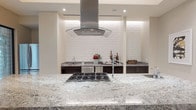 This single-story space showcases a thoughtfully designed modern kitchen that combines clean lines with high-end finishes. The centerpiece is a large granite island with striking gray-and-white veining, equipped with a professional-grade stainless steel gas cooktop, making it as functional as it is visually impressive.

A bold stainless steel island range hood with a curved glass canopy serves as a striking architectural focal point, suspended above the cooktop and drawing the eye upward. The white subway tile backsplash extends across the back wall, framed by recessed ambient lighting that casts a warm, soft glow along the upper perimeter, adding depth and a refined atmosphere to the space.

Dark espresso cabinetry lines the back wall, offering a rich contrast against the light granite countertops and white tile. Additional counter space is visible to the sides, providing generous prep and storage areas. Recessed ceiling lighting throughout ensures the kitchen is well-illuminated, while the neutral cream-toned walls contribute to a bright, open, and cohesive aesthetic overall.