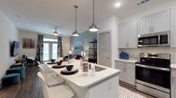 This single-floor residence offers a thoughtfully designed open-concept layout that seamlessly connects the kitchen, dining, and living areas. The kitchen is a standout feature, showcasing crisp white cabinetry, light-toned countertops, and a large central island with seating — ideal for casual dining or entertaining. A classic subway tile backsplash adds texture, while stainless steel appliances contribute a sleek, modern edge. Three elegant pendant lights hang above the island, casting warm, focused illumination that complements the recessed ceiling lighting throughout. The living area is visible from the kitchen, creating a cohesive, airy atmosphere enhanced by large windows dressed with patterned curtains that invite generous natural light. A ceiling fan adds both function and style to the living space. The flooring transitions smoothly from tile to wood-look surfaces, grounding the open layout with warmth and continuity. Soft, neutral tones in blue-grey and white define the color palette, creating a clean and inviting ambiance. The overall design balances contemporary aesthetics with everyday practicality, making it a comfortable and visually appealing living space.