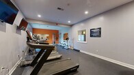 Located at 48 Bavarian Way in Altamont, NY, this dedicated fitness center offers a comprehensive workout environment housed within a single, open-plan exercise room. The space is finished with durable dark rubber flooring throughout, providing a functional and low-maintenance surface suited for high-activity use.

The room is thoughtfully organized to accommodate a wide variety of training styles. Cardio equipment includes treadmills, an elliptical trainer, a stationary upright bike, and a rowing machine, while strength training is supported by a dumbbell rack, a weight bench, and a cable/resistance station. A wall-mounted speed bag platform adds a striking boxing-oriented feature to one corner of the room.

Recessed lighting illuminates the space evenly, complemented by natural light from windows that offer views of the surrounding exterior. A bold orange accent wall with large mirrors anchors the strength training area, adding visual energy and depth to the room. Wall-mounted flat-screen televisions are positioned for use during cardio sessions. Motivational framed artwork is displayed throughout, reinforcing an active and goal-oriented atmosphere.

The neutral gray walls and clean white trim provide a polished backdrop, while the open layout ensures ample floor space for stretching and floor exercises. This fitness center delivers a well-rounded facility suited to a diverse range of fitness goals and routines.