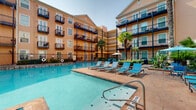 This outdoor pool amenity space in New Orleans, LA, offers a well-appointed, resort-inspired retreat set within a multi-story courtyard building. The centerpiece is a generously sized swimming pool with clear, blue water, surrounded by a tiled deck lined with blue lounge chairs, providing ample space for sunbathing and relaxation. Lush palm trees and tropical plantings are interspersed throughout, enhancing the warm, leisure-focused atmosphere.

A standout architectural feature is the covered colonnade running alongside the pool, defined by a series of elegant arched openings that frame views of the water and surrounding greenery. This shaded corridor is furnished with blue high-top tables and chairs as well as lounge seating arrangements, making it a comfortable gathering space even during New Orleans' warmer months. Ceiling fans are installed overhead to promote air circulation throughout the covered area.

Adjacent to the colonnade is an enclosed lounge alcove, furnished with modern blue cushioned seating and a coffee table, accented by decorative wall art. A vending machine is conveniently located nearby. Tile flooring runs throughout the covered zones, while the multi-story building facades featuring private balconies enclose the courtyard, creating an intimate and sheltered atmosphere.
