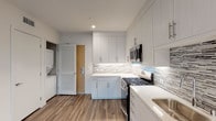 This single-floor unit showcases a contemporary design with wood-style flooring and a neutral color palette carried throughout. The open-concept living and kitchen area is bright and airy, with multiple windows allowing natural light to fill the space and a glass door leading to a private balcony with outdoor views.

The kitchen is well-appointed with floor-to-ceiling white shaker cabinetry, under-cabinet lighting, a textured mosaic tile backsplash, white quartz countertops, and a full suite of stainless steel appliances including a gas range, over-the-range microwave, refrigerator, and dishwasher. A stacked washer and dryer unit is conveniently housed within the kitchen area.

The bedrooms are generously sized with clean white walls, wood-style flooring, and ample natural light. The primary bedroom includes access to an en-suite bathroom and a walk-in closet with built-in shelving. The bathrooms are elegantly finished with marble-look porcelain tile surrounds on the soaking tubs, mosaic tile flooring, white vanities with quartz countertops, and built-in open shelving, creating a clean, spa-like aesthetic throughout.